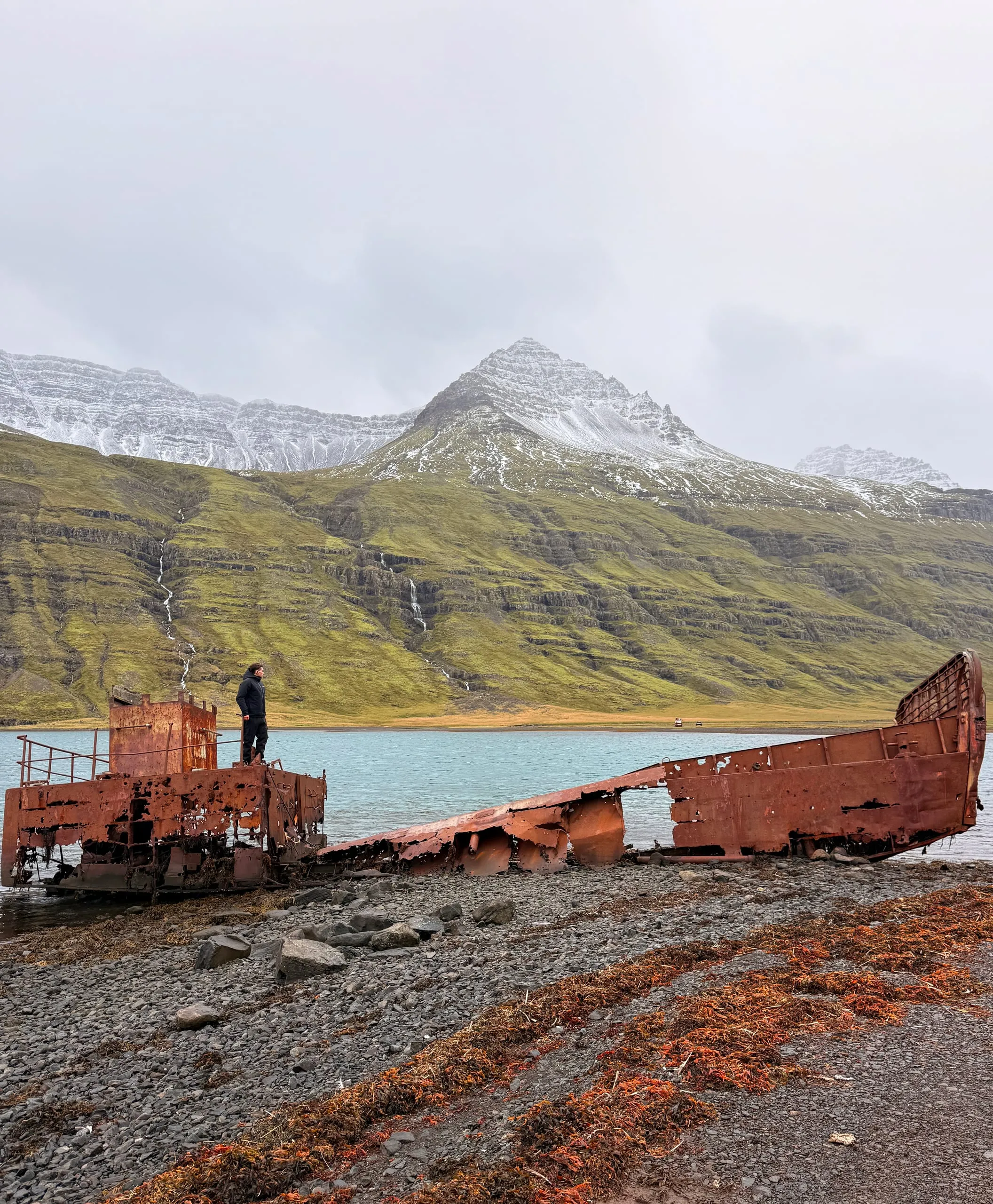 A rusted wartime landing craft beached at the head of an Icelandic fjord, snow-streaked mountains and a waterfall behind it