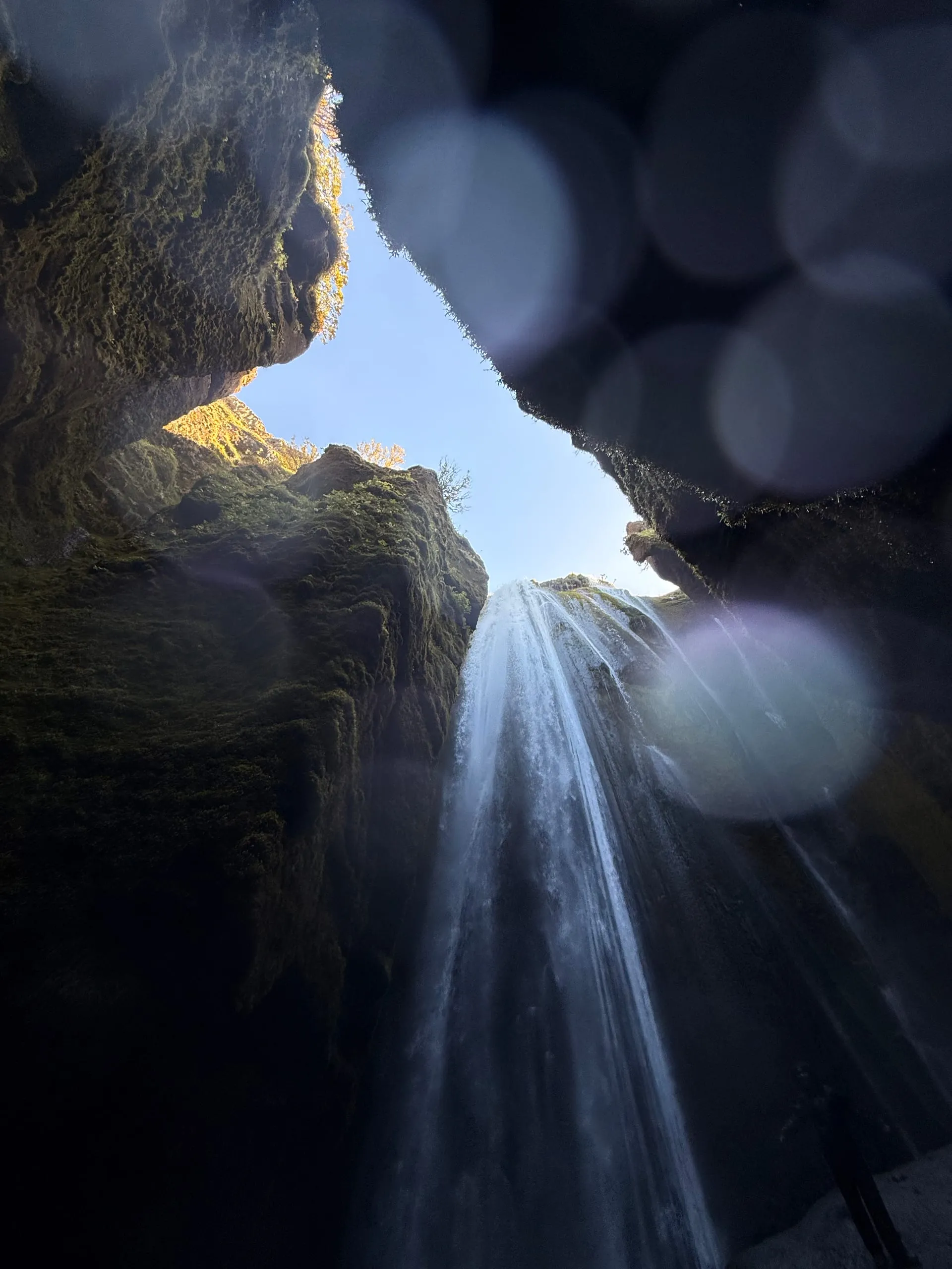 A waterfall seen from inside a narrow mossy cleft, falling toward the camera with a slot of blue sky visible between the overhanging walls