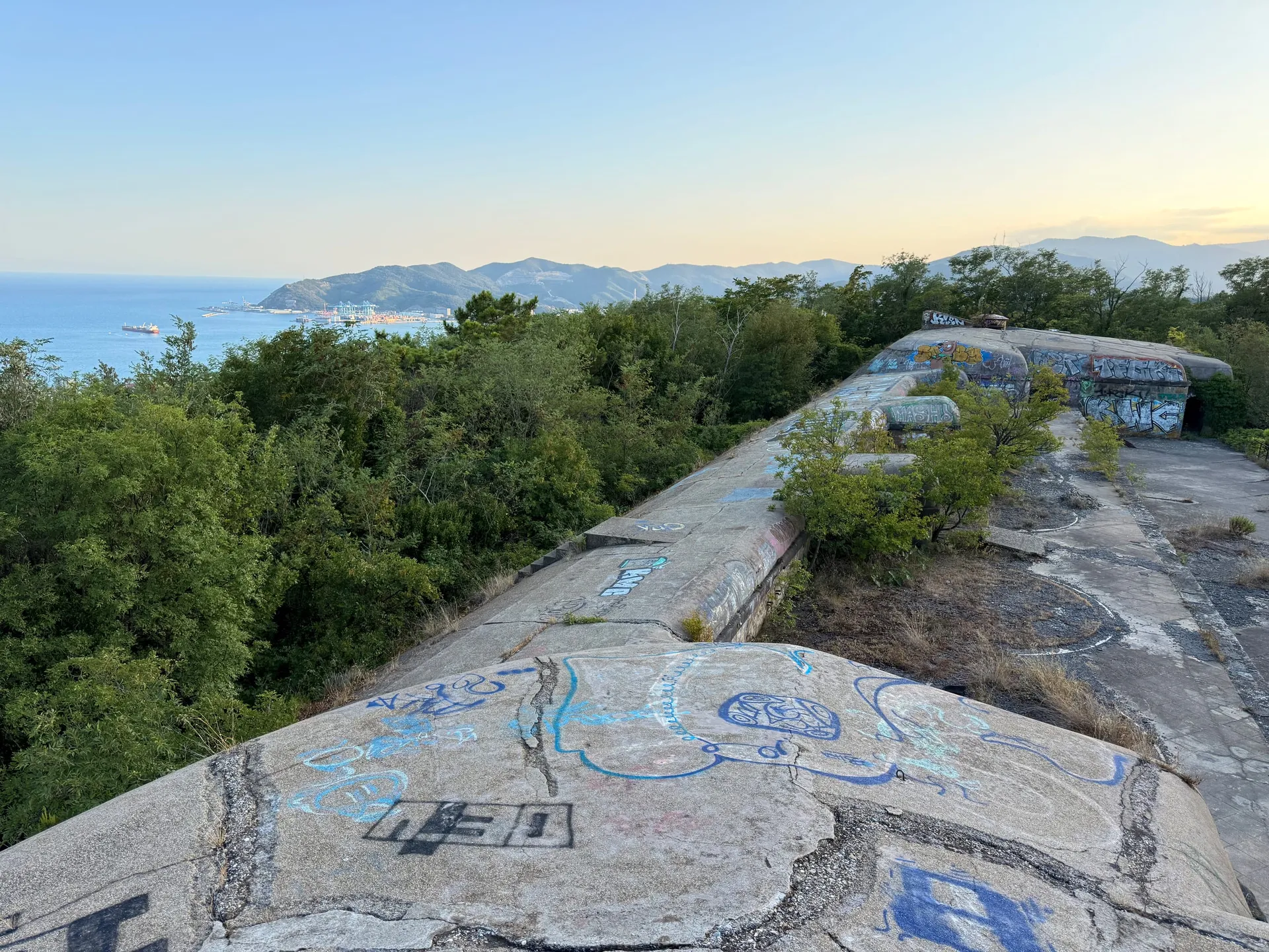 The concrete roof of a coastal gun battery above Savona — cracked, graffitied slabs running toward armoured cupolas, with the Ligurian Sea, the port cranes and blue headlands beyond in evening light