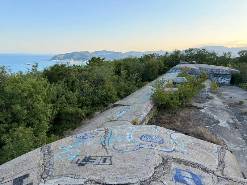 The concrete roof of a coastal gun battery above Savona — cracked, graffitied slabs running toward armoured cupolas, with the Ligurian Sea, the port cranes and blue headlands beyond in evening light