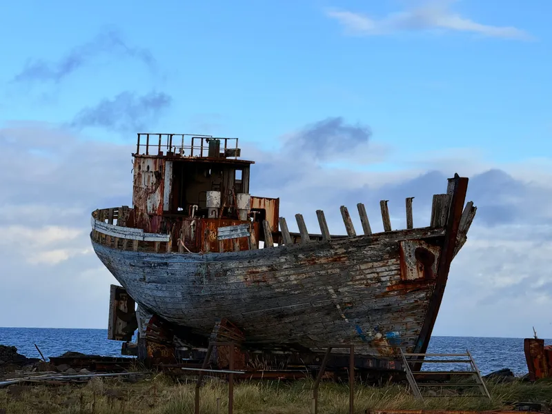 A wooden herring boat propped upright on a steel cradle on the Icelandic shore, its planking weathered silver, bare frames standing above the sheer and a rusted wheelhouse aft, the sea and morning sky behind