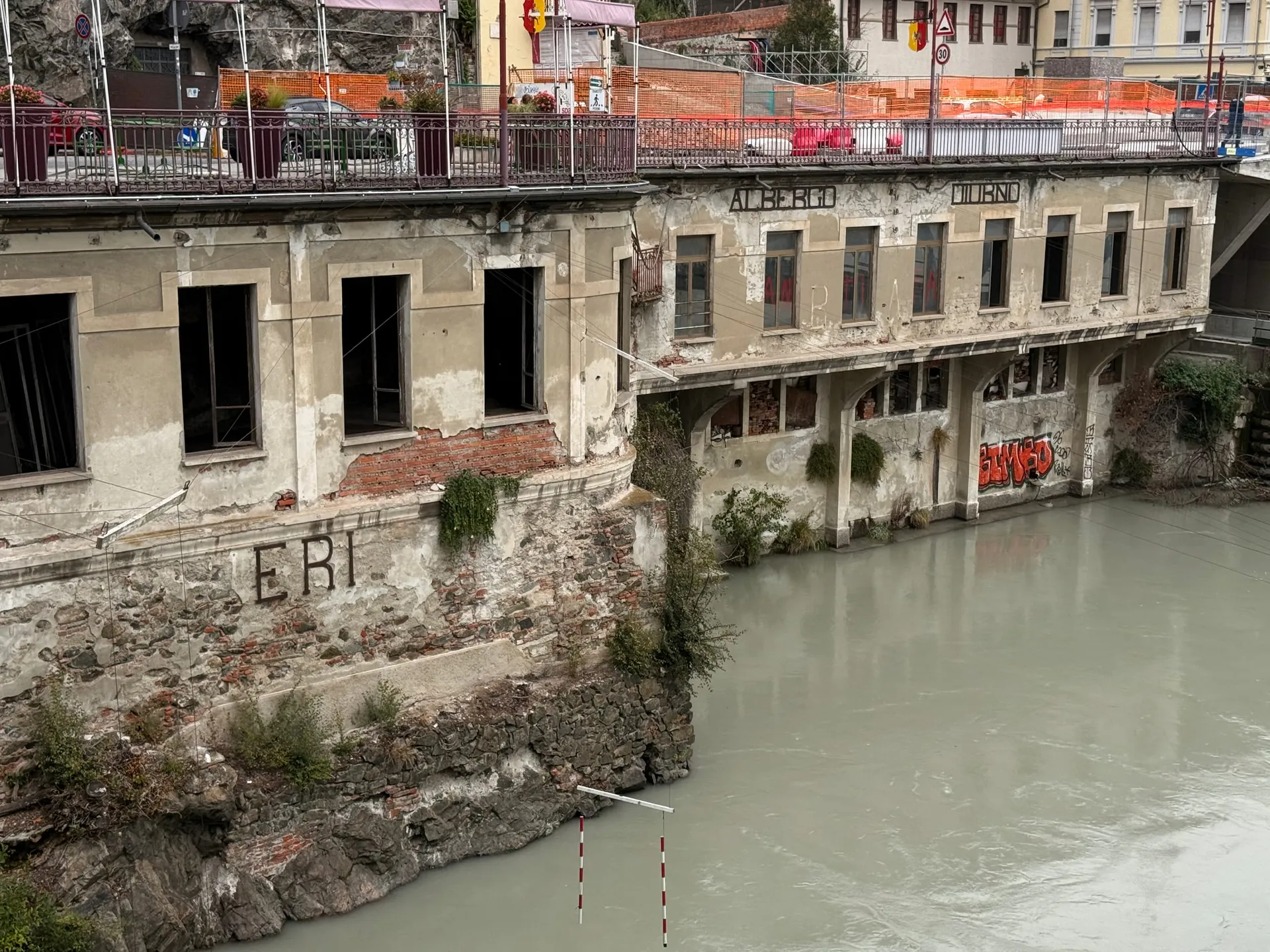 A long, low derelict building standing on bare rock at the edge of a pale green river in Ivrea, its stripped windows in a row and the ghost lettering "ALBERGO DIURNO" still legible across the facade, with the street railing and traffic passing directly above the roof