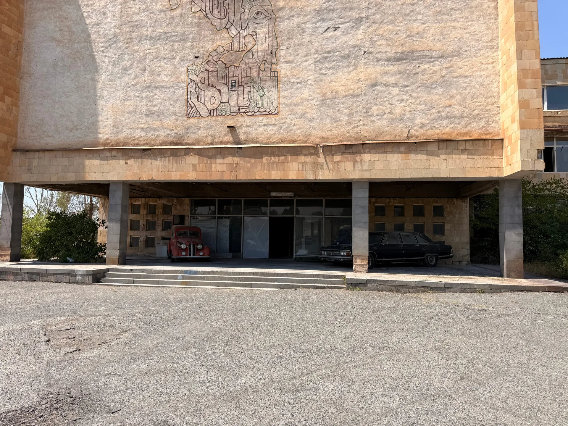 A tuff-faced studio block standing on concrete piers with a large incised line drawing of a face and a city across its upper wall, two old cars parked in the shaded undercroft below