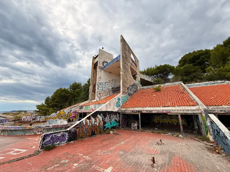 An abandoned concrete resort building above the Adriatic, a tall pierced fin standing clear of pantile roofs over a red-brick terrace, every surface covered in graffiti
