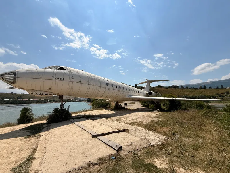 A sun-bleached Soviet Tupolev Tu-134A airliner parked on a dirt bluff above a turquoise reservoir, its glazed navigator's nose out over the water and cables of festoon lighting running the length of the fuselage