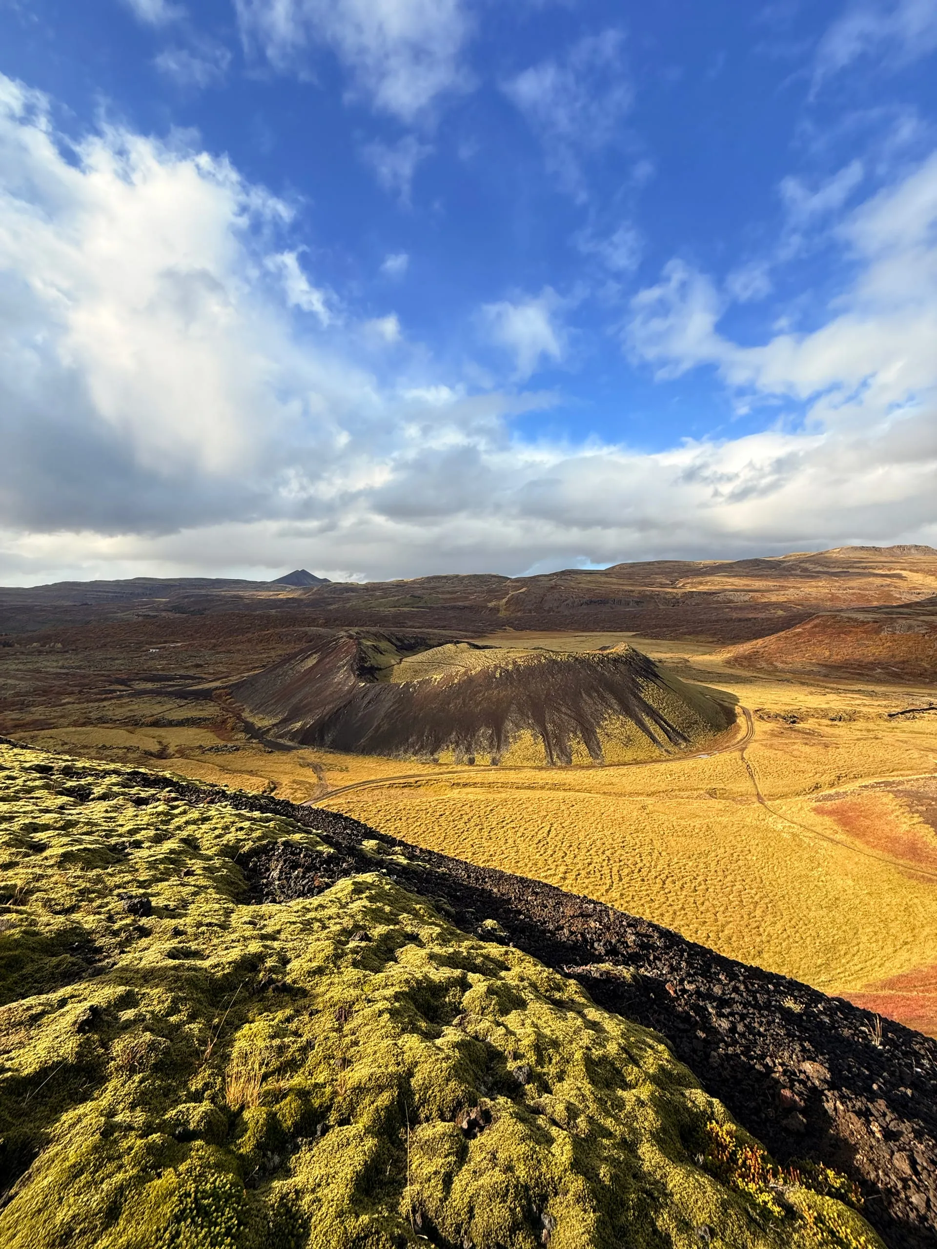 A dark volcanic crater rising from a yellow-gold plain, seen from a neighbouring rim covered in bright green moss, under a big sky of broken cloud