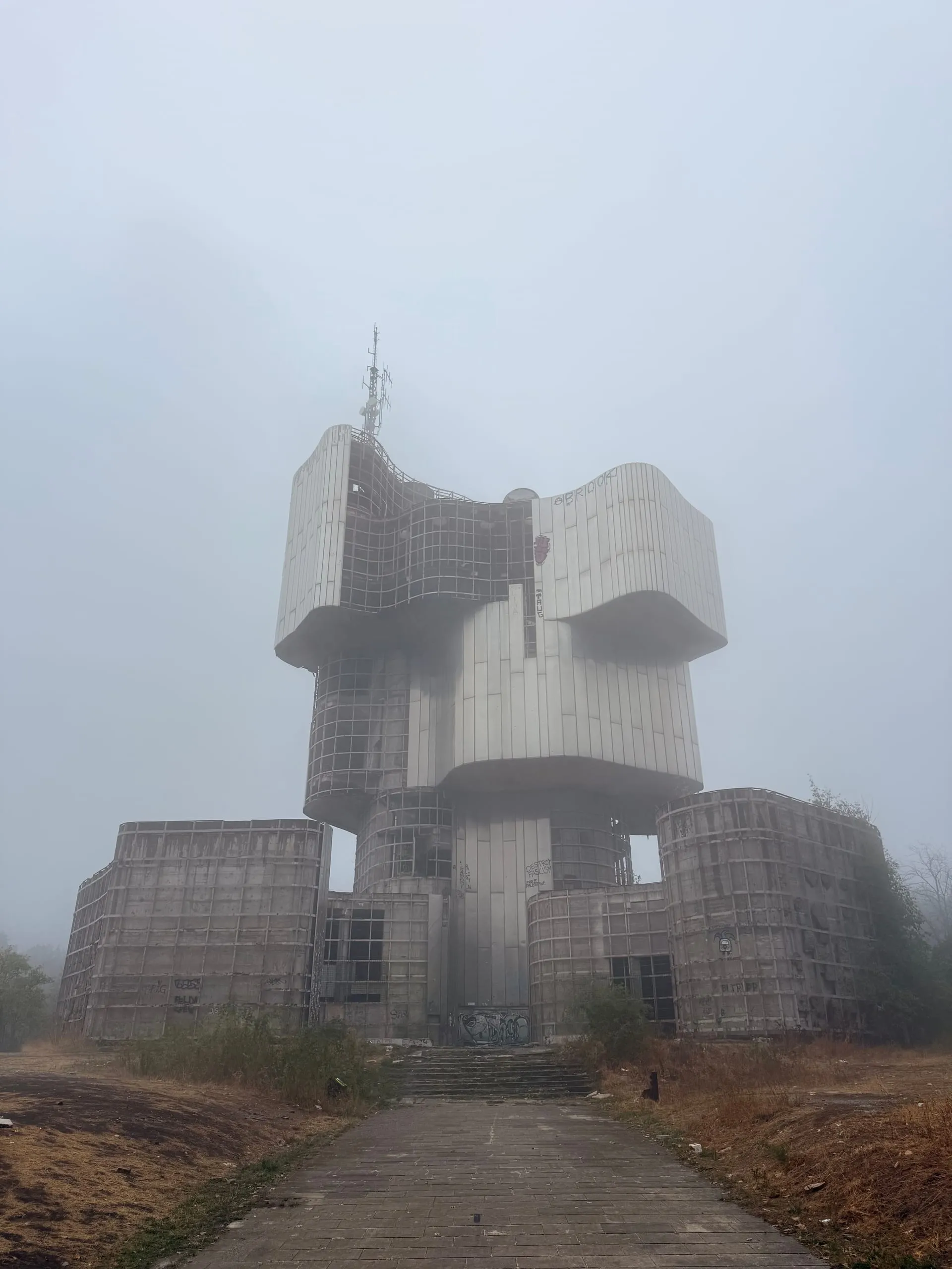 A tall abandoned memorial of stacked, swelling forms at the end of a paved avenue, half dissolved in thick fog, a mast rising from its top