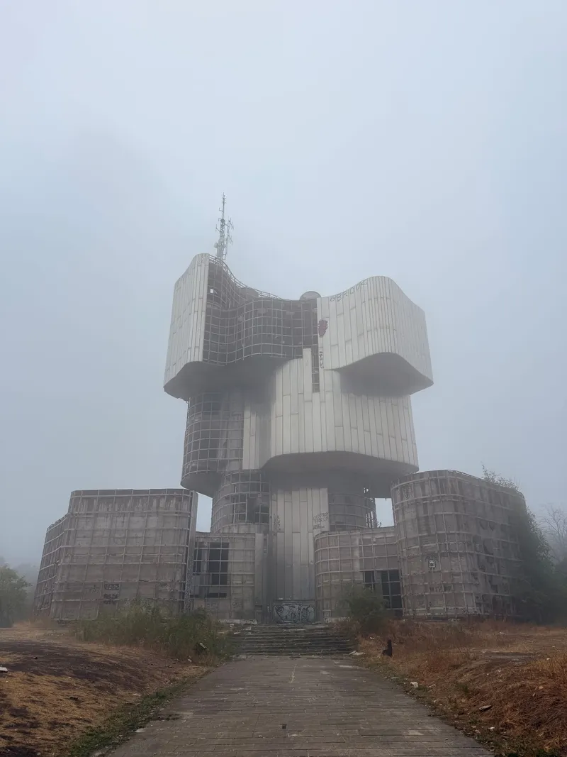 A tall abandoned memorial of stacked, swelling forms at the end of a paved avenue, half dissolved in thick fog, a mast rising from its top