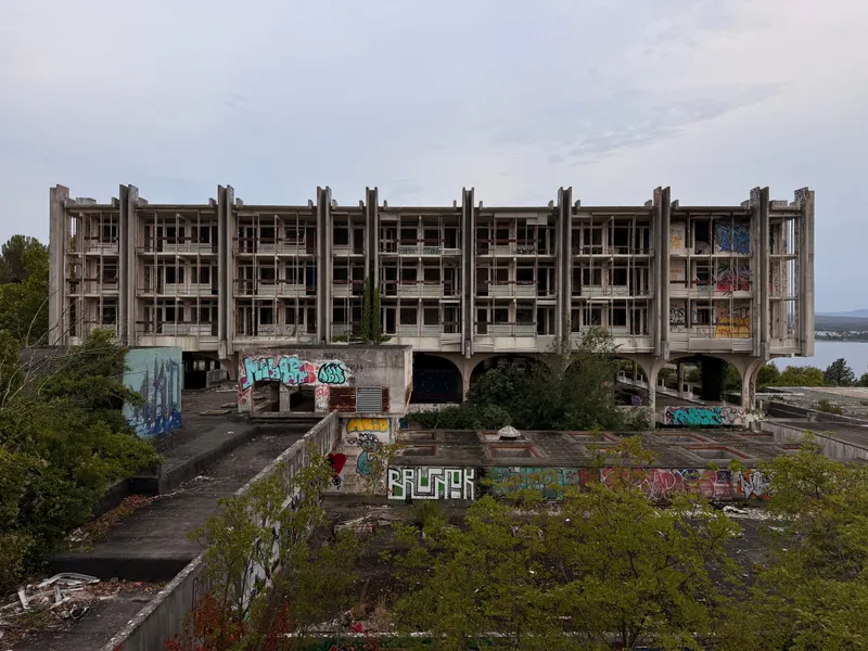 A long four-storey abandoned concrete hotel with a deep grid of loggias across its whole face, graffiti along the base, scrub growing over the terrace in front of it