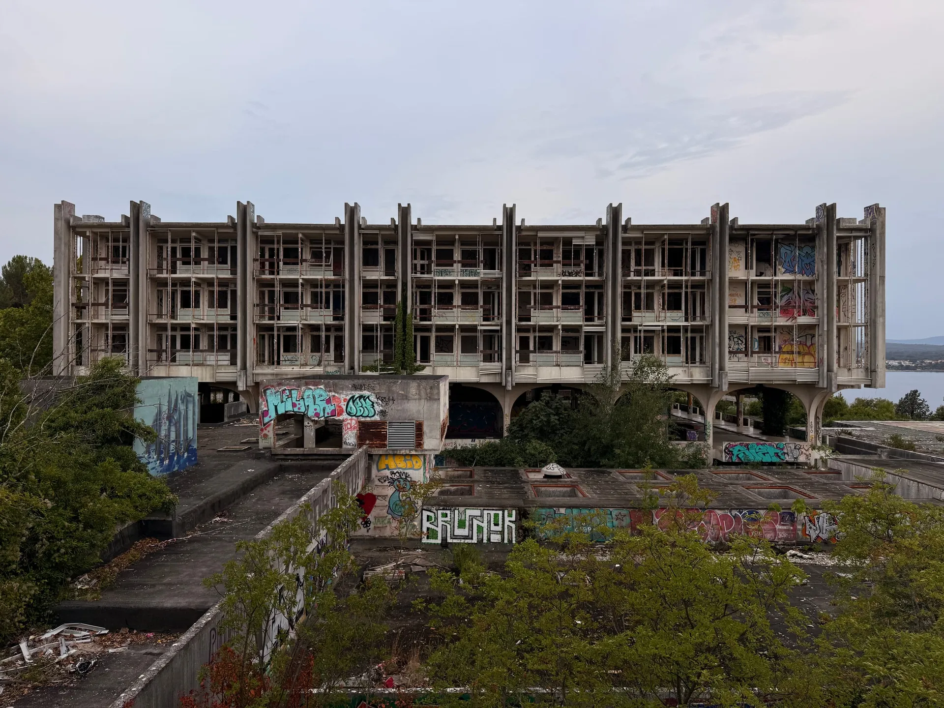 A long four-storey abandoned concrete hotel with a deep grid of loggias across its whole face, graffiti along the base, scrub growing over the terrace in front of it