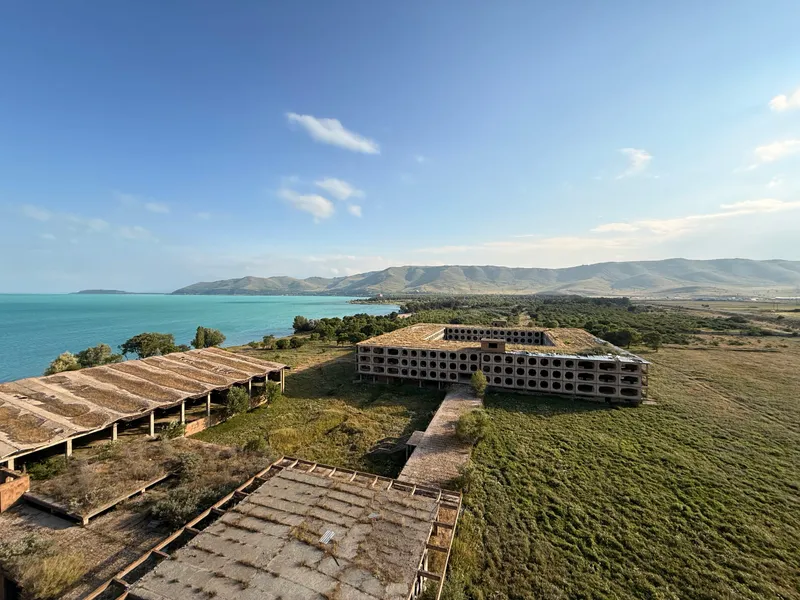 An unfinished lakeside resort complex seen from above — long bare-roofed wings and a block with a facade of stacked concrete rings, the turquoise water of Lake Sevan and bare summer hills beyond