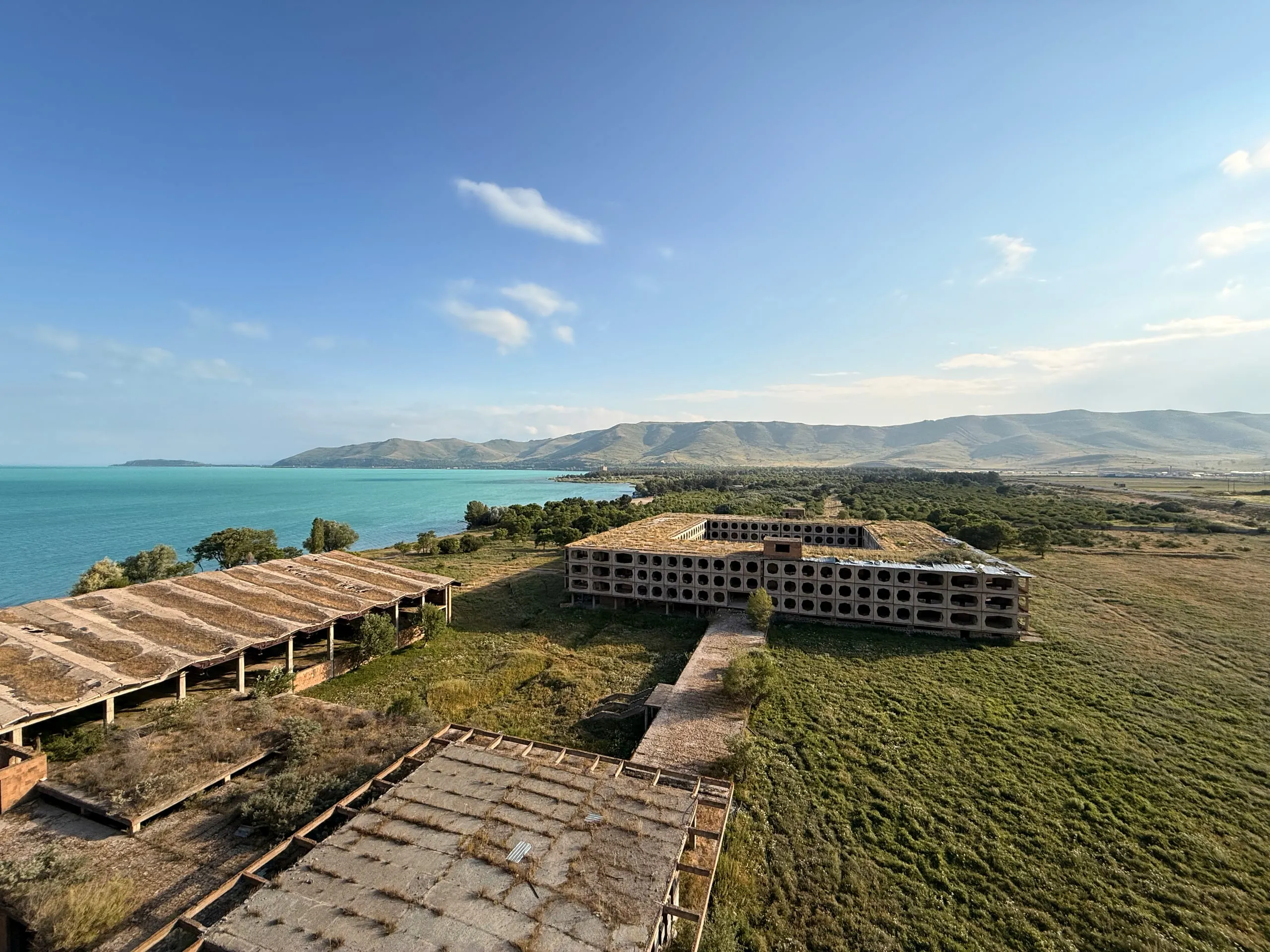 An unfinished lakeside resort complex seen from above — long bare-roofed wings and a block with a facade of stacked concrete rings, the turquoise water of Lake Sevan and bare summer hills beyond