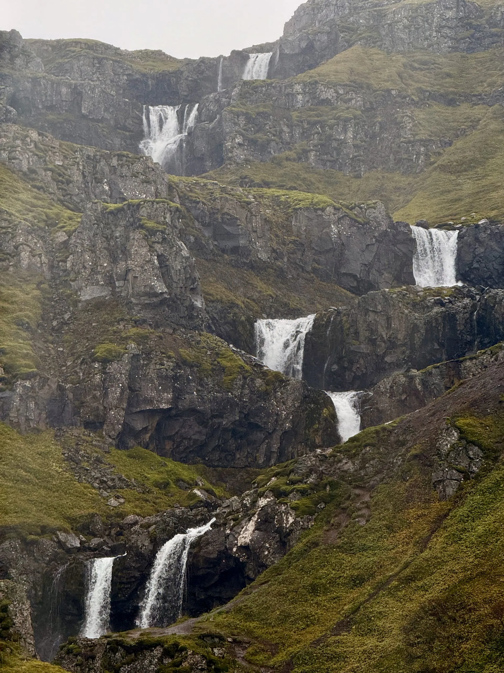 A stepped cascade of many waterfalls coming down a steep green-and-grey valley wall in low cloud