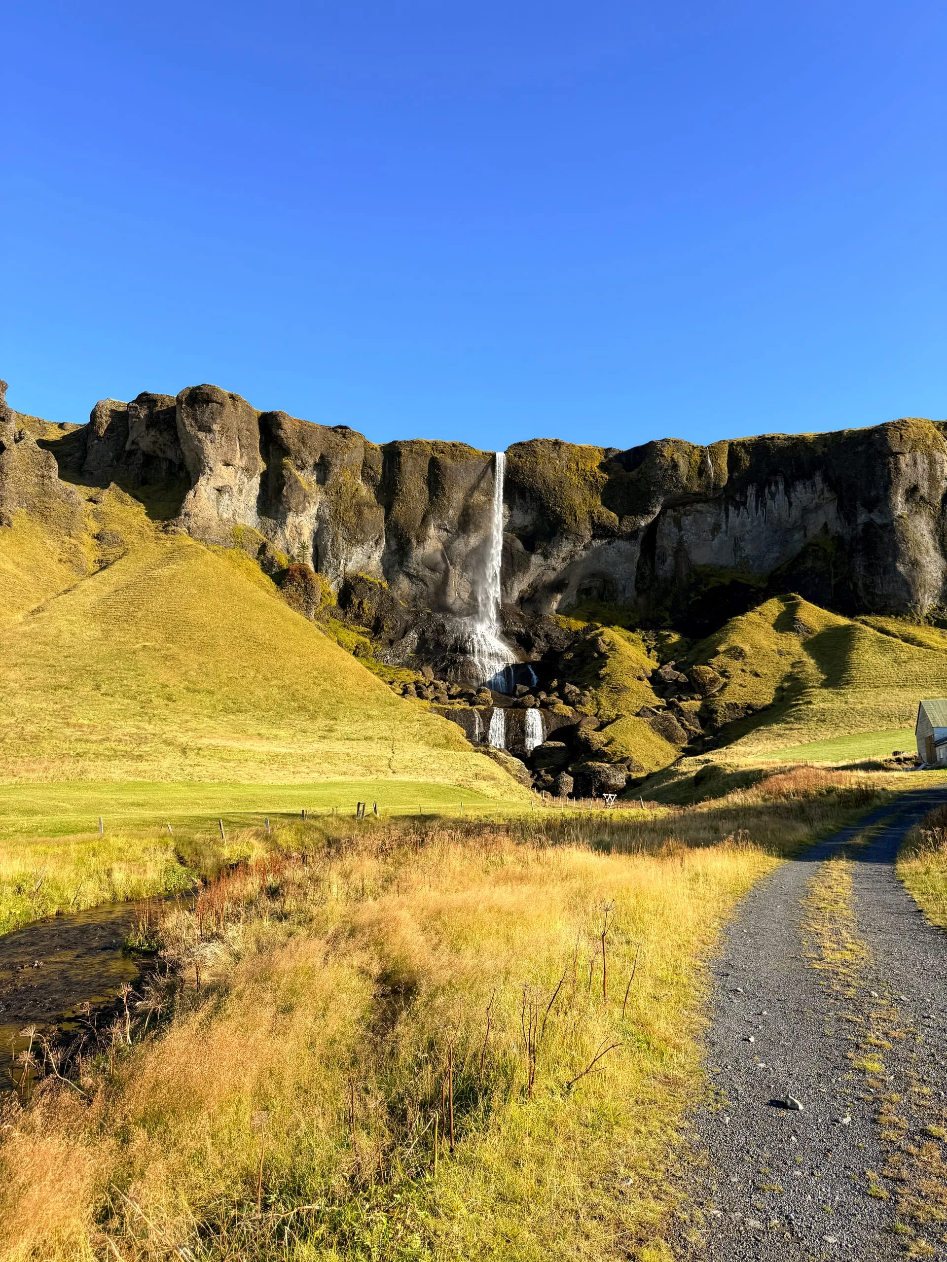 A tall thin waterfall dropping off a long dark cliff onto a green field, with a gravel track running toward it through golden autumn grass