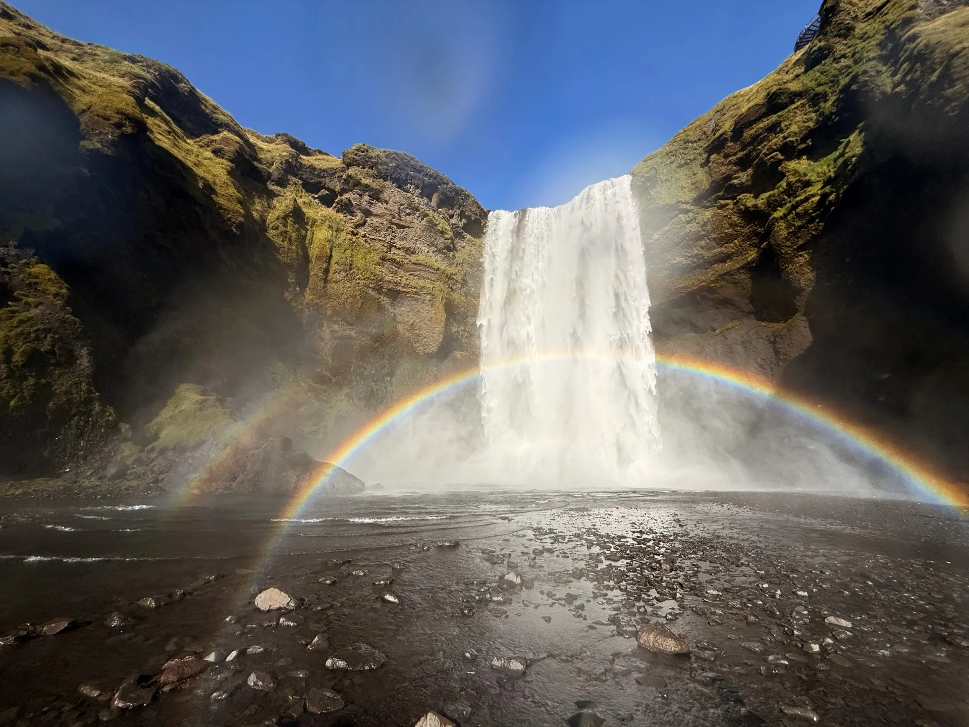 A wide sixty-metre waterfall falling from a green-and-black cliff into a gravel plain, with a complete rainbow arching across the spray in front of it under a deep blue sky