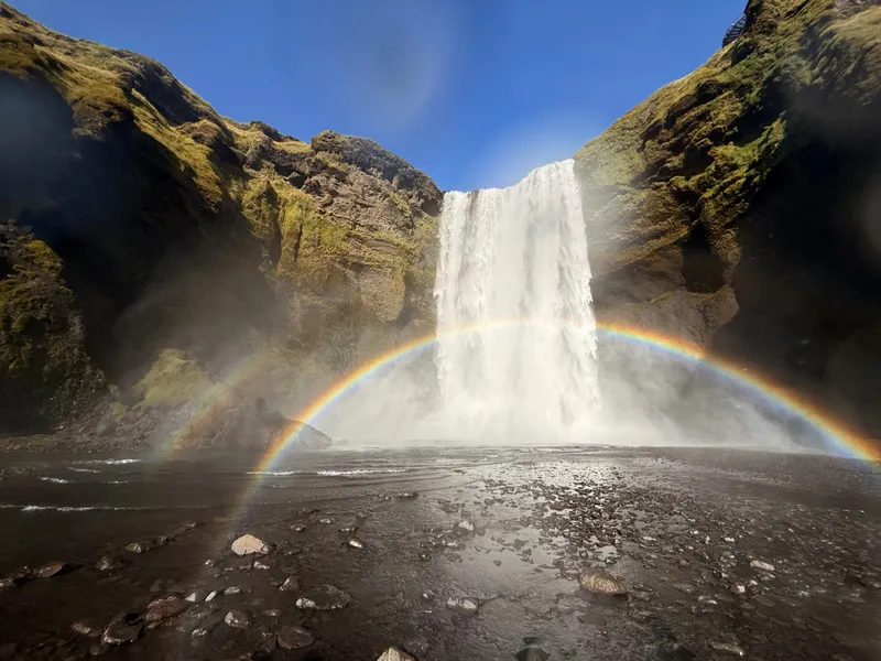 A wide sixty-metre waterfall falling from a green-and-black cliff into a gravel plain, with a complete rainbow arching across the spray in front of it under a deep blue sky