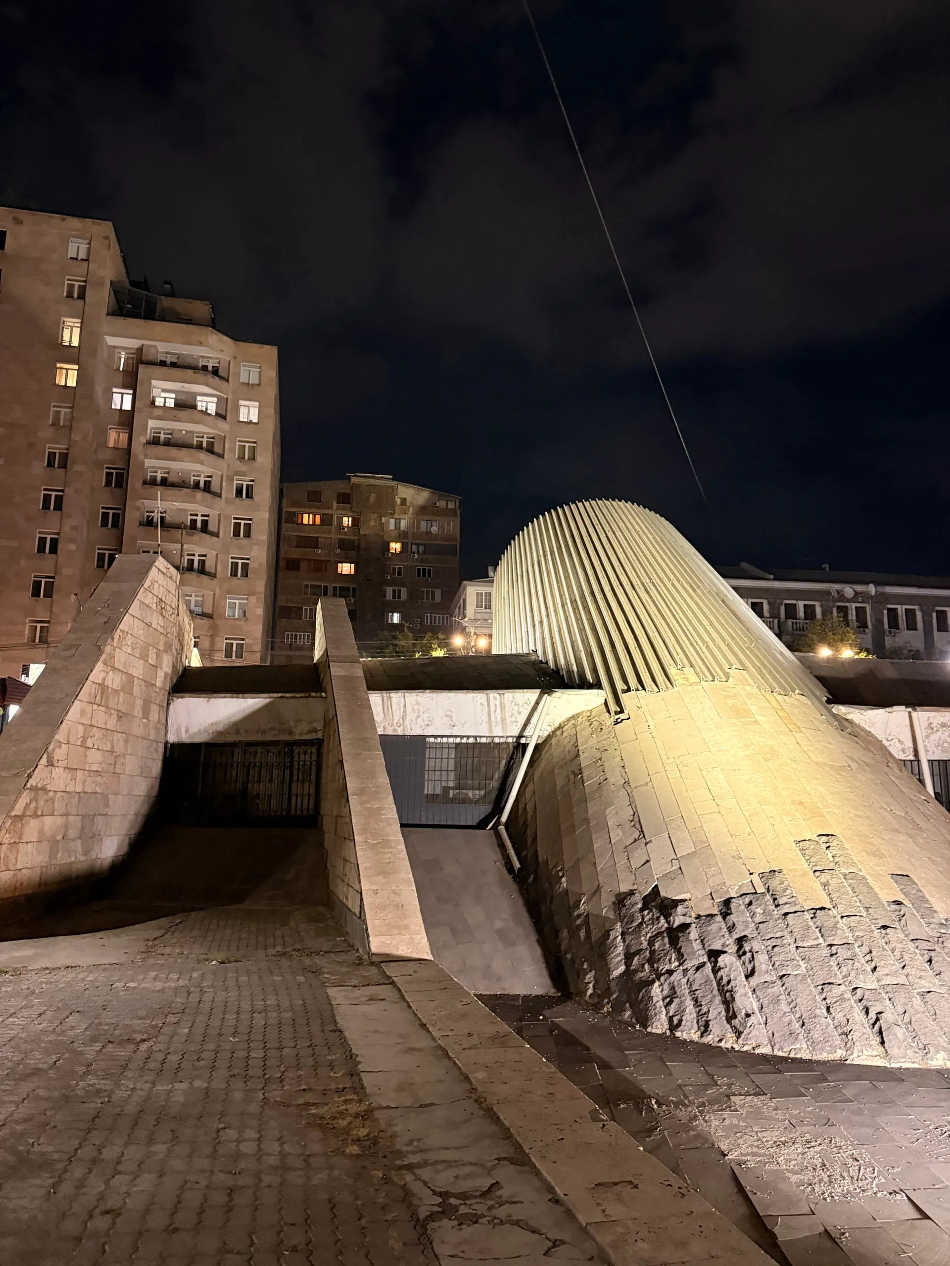 A ribbed metal shell breaking out of the ground at an angle above a concrete ramp descending into a metro entrance at night, lit warm, with Soviet apartment blocks behind it