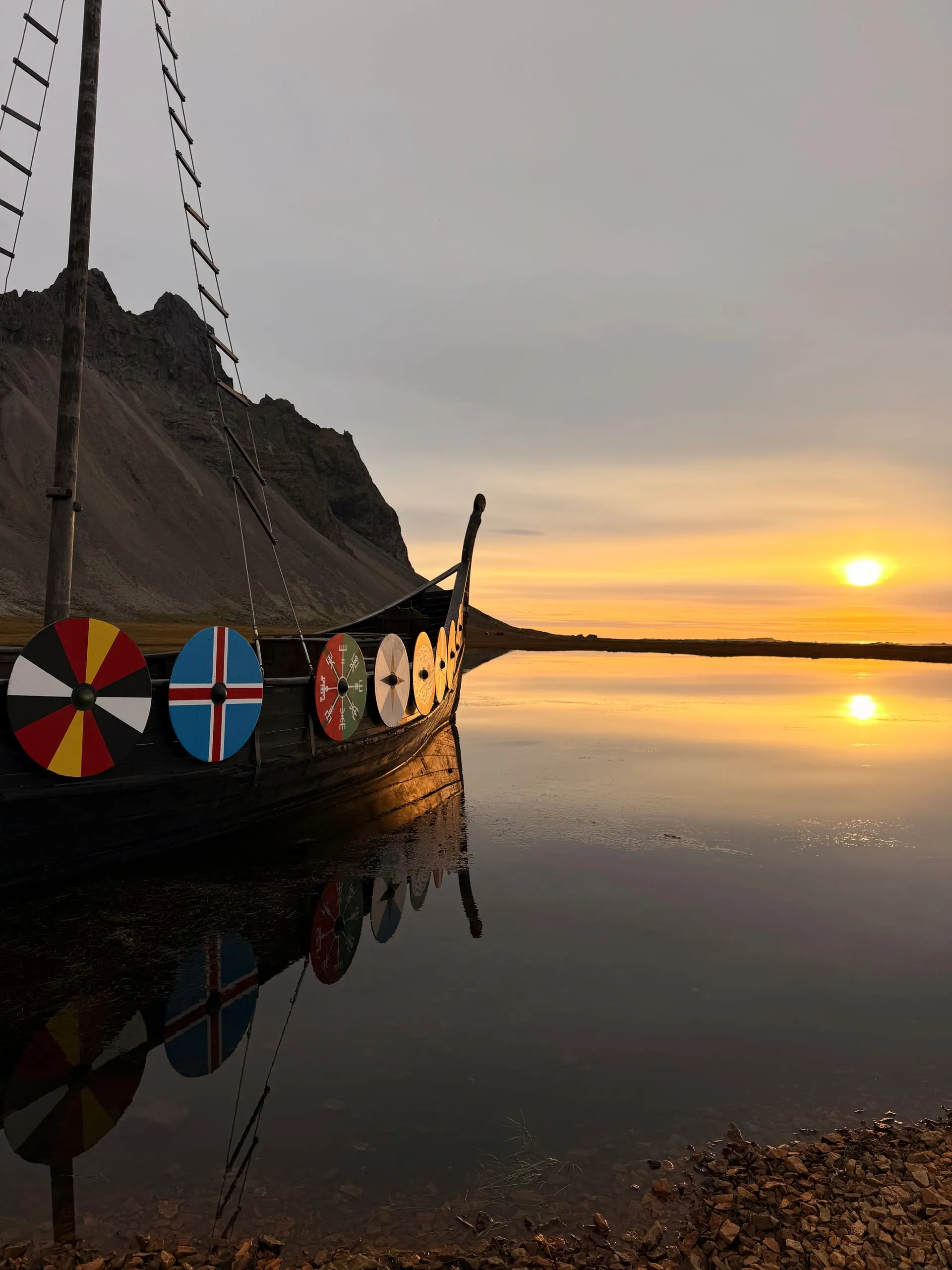 The bow of a wooden longship with painted round shields along its side, drawn up on a flat tidal shore at sunrise, the dark peaks of Vestrahorn rising behind it