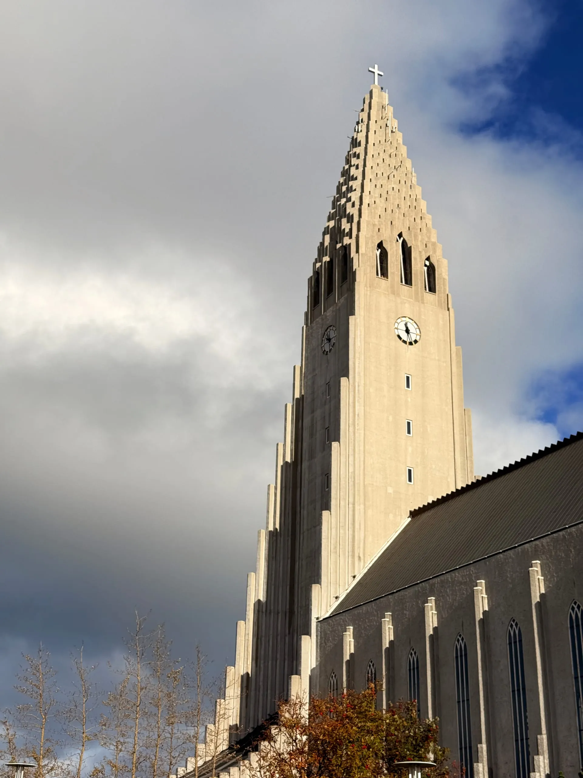 The concrete tower and stepped west front of Hallgrímskirkja seen from below at an angle, lit by low sun against a dark storm cloud