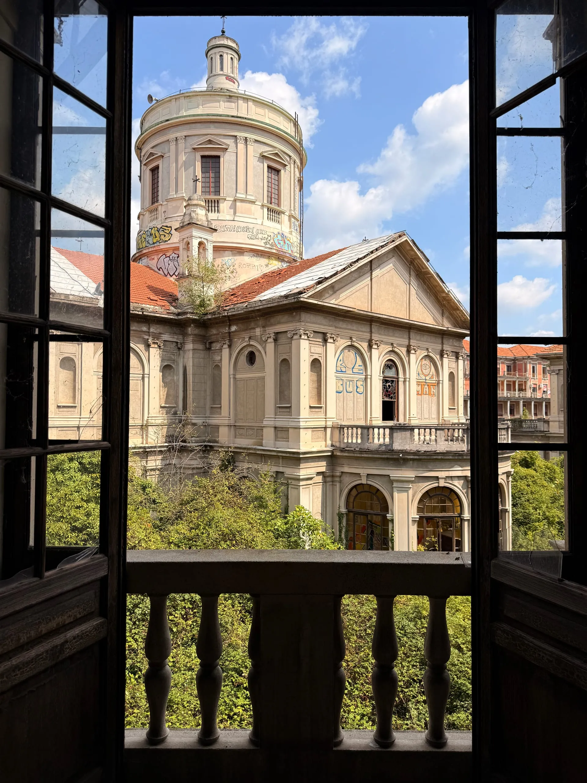 The bone-coloured dome of an abandoned sanatorium chapel, framed through an open window with a stone balustrade and an overgrown courtyard below