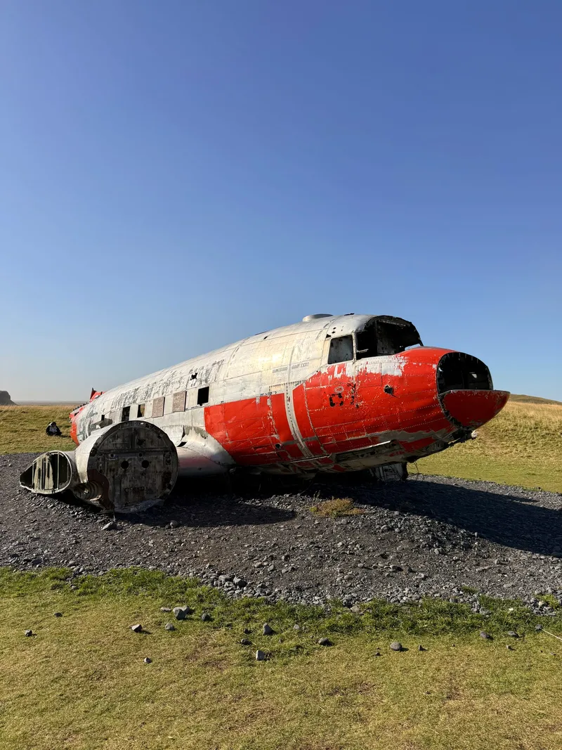 A wingless US Navy DC-3, bare aluminium with a bright red nose and belly band, standing on a gravel bed in a green Icelandic meadow under a clear blue sky