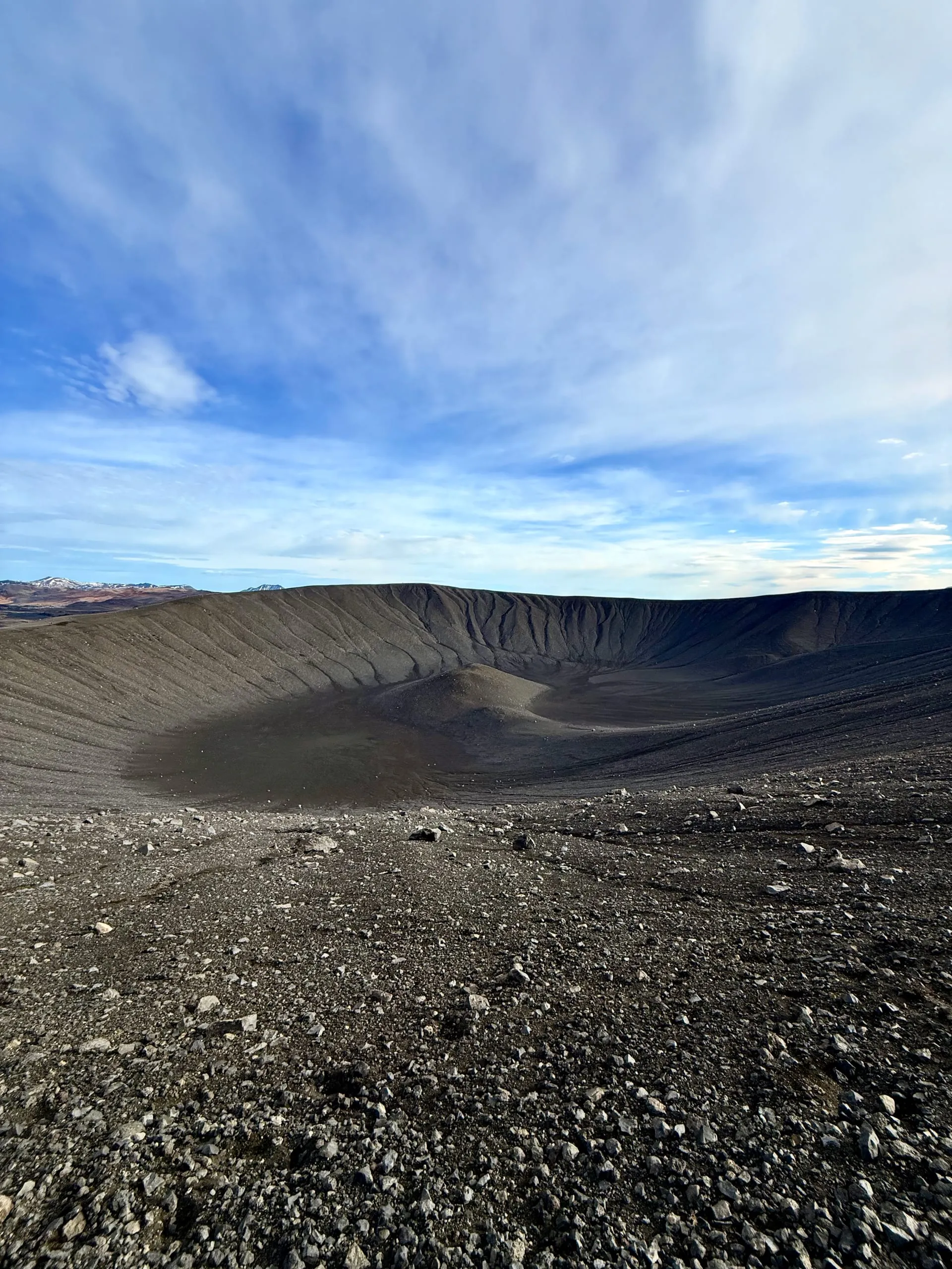 The interior of a vast circular volcanic crater of dark grey ash and gravel, seen from the rim, with a low mound at its centre and mountains on the far horizon
