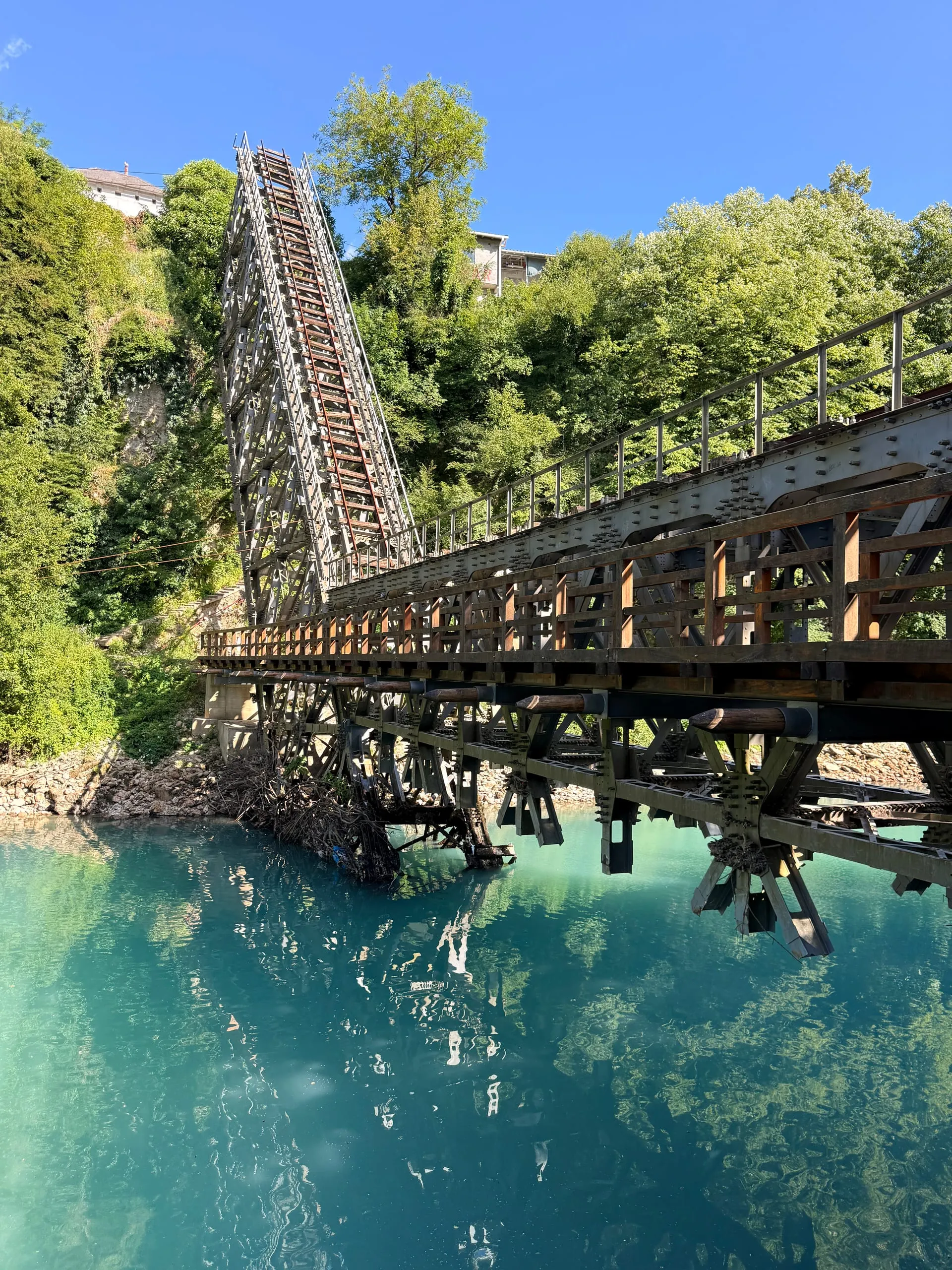 The buckled steel truss of a broken railway bridge dropping side-on into the emerald-green Neretva river
