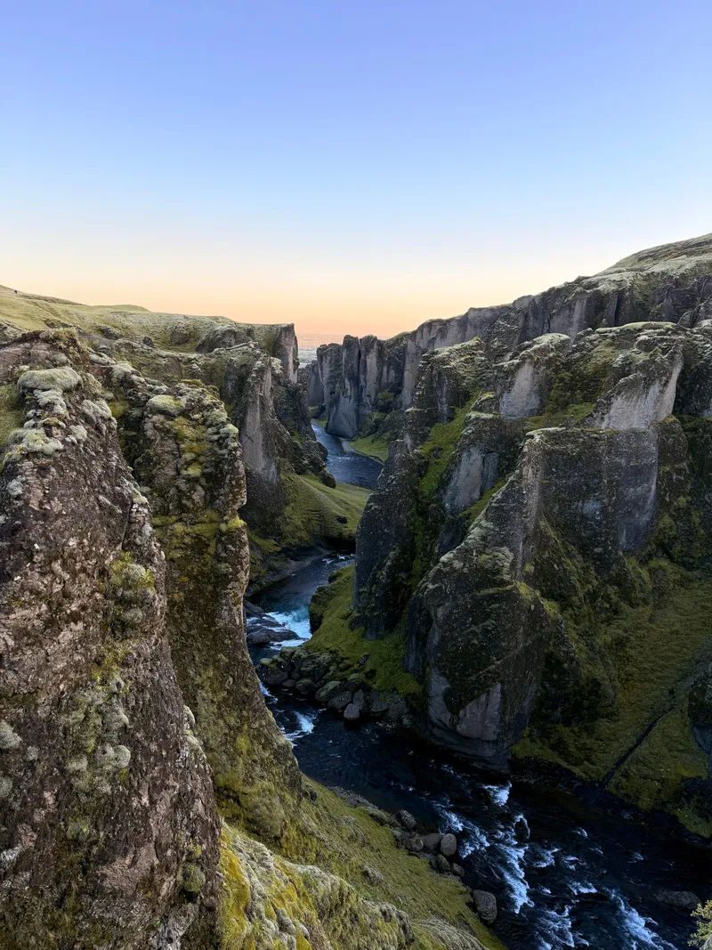A deep serpentine canyon of moss-covered rock towers at dusk, a small river winding along its floor, the sky going orange at the horizon