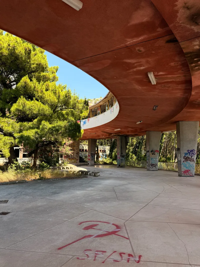 A circular abandoned concrete sanatorium raised on columns, hidden among coastal pines above the Adriatic