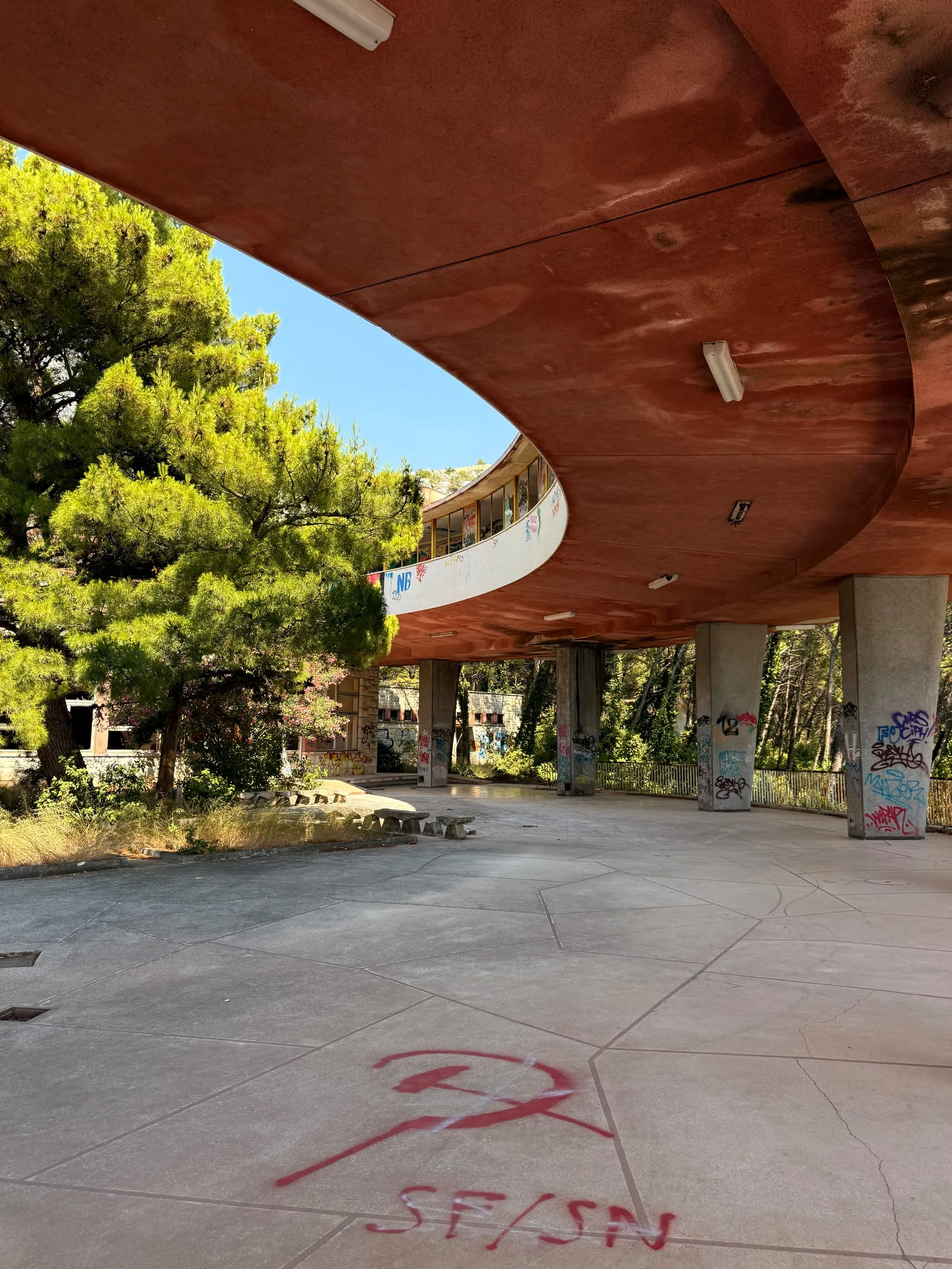 A circular abandoned concrete sanatorium raised on columns, hidden among coastal pines above the Adriatic