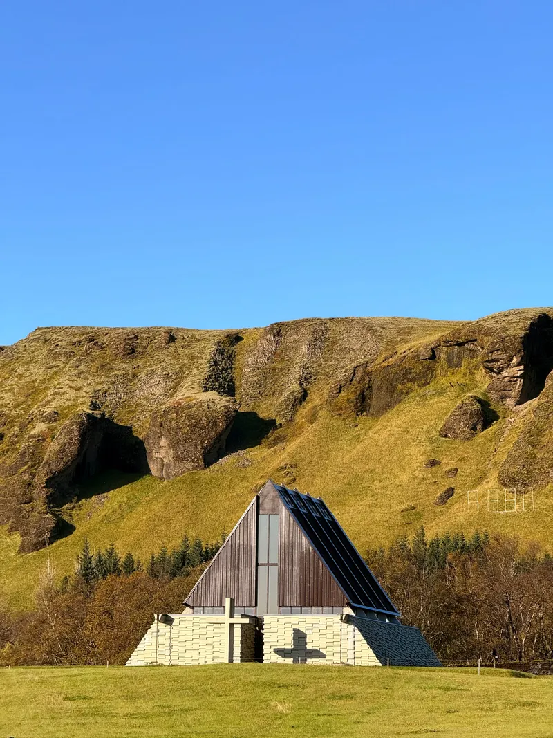 A dark timber A-frame chapel with a steep slate roof running to the ground, standing on a pale stone base in a green field below a cliff of columnar rock