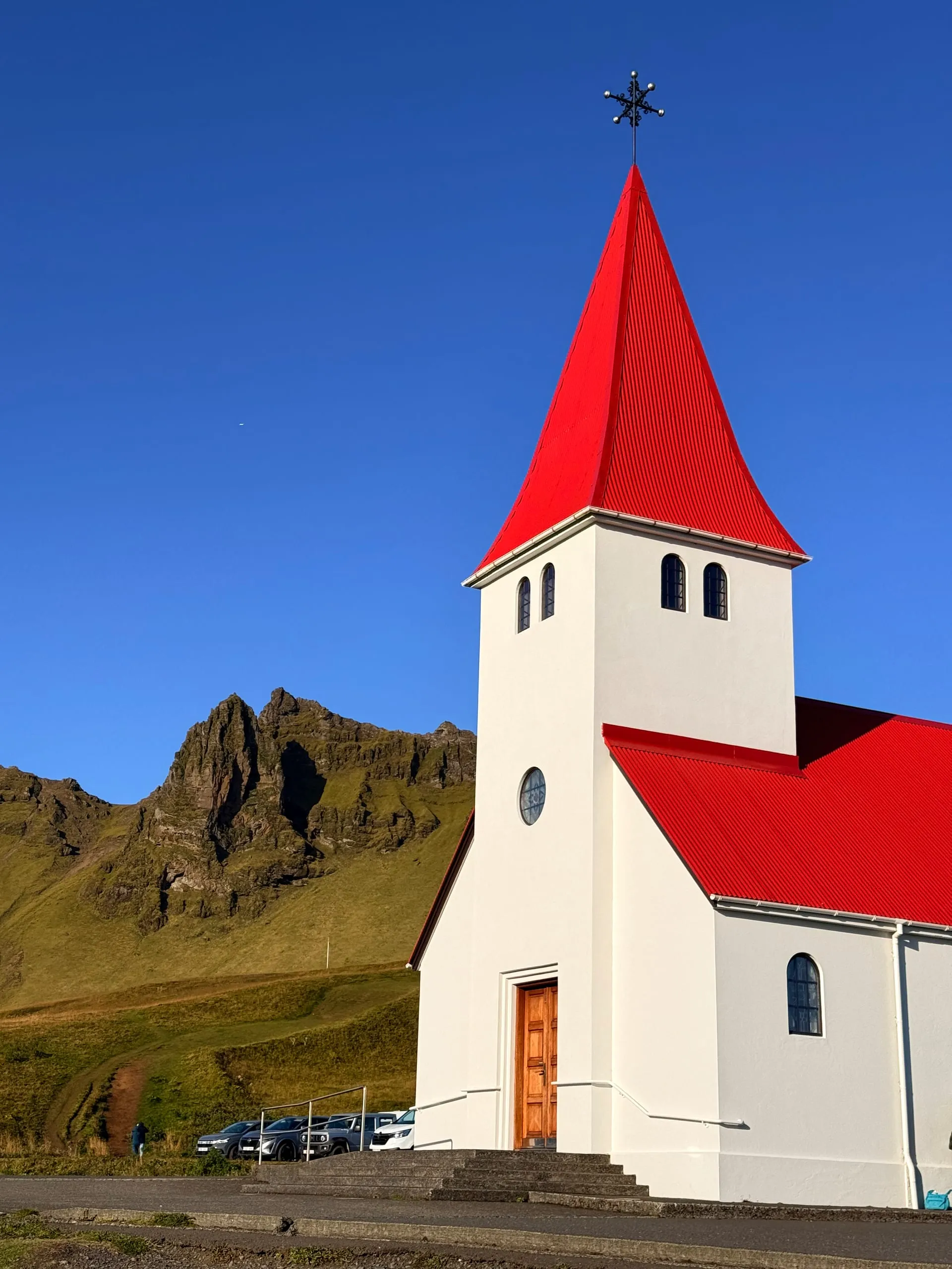 A white church with a steep red roof and a small tower standing on a green slope, with jagged dark crags rising behind it under a clear sky