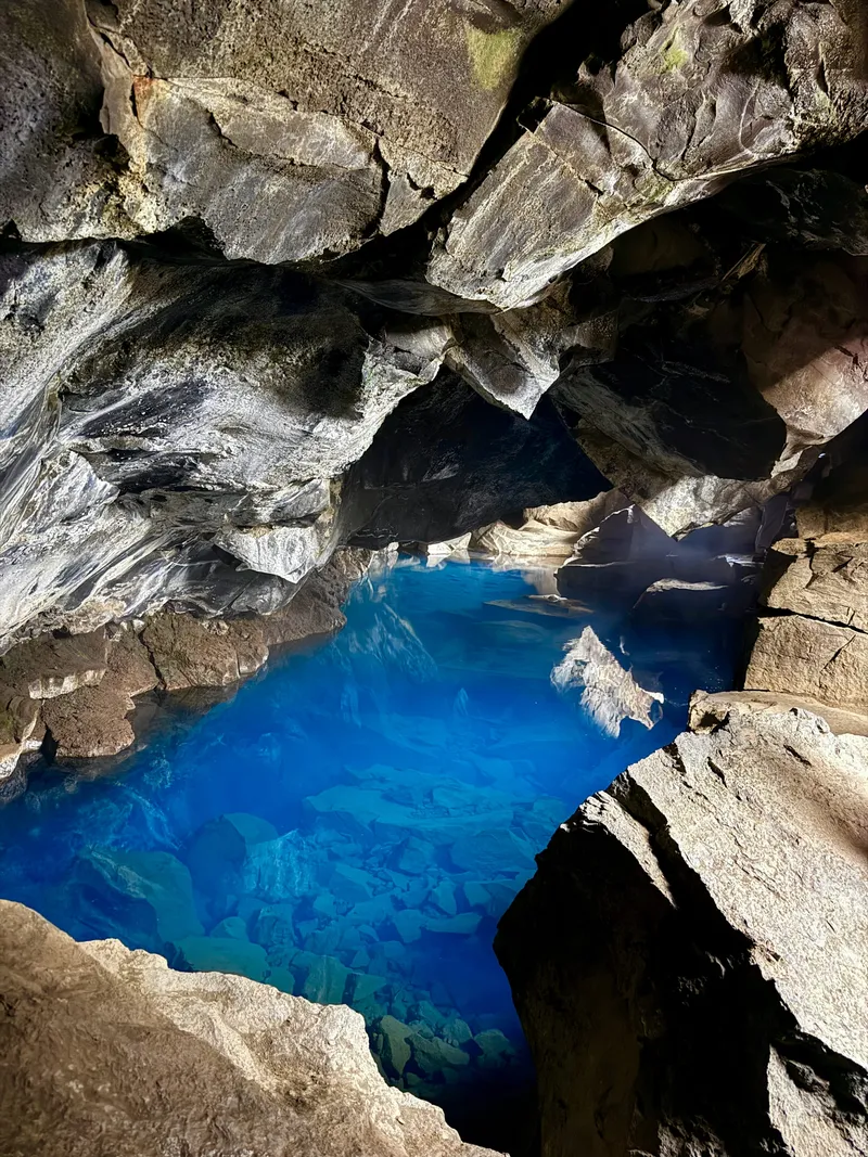 Brilliant blue geothermal water in a lava cave, lit by daylight coming through a crack in the rock overhead, with jagged basalt walls closing in around it