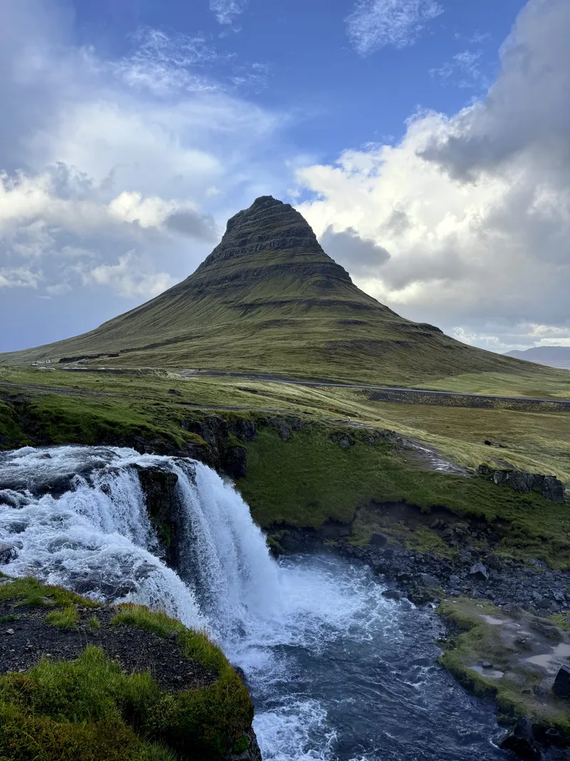 A steep isolated conical mountain across a green plain, with a broad three-step waterfall in the foreground under a sky of broken cloud