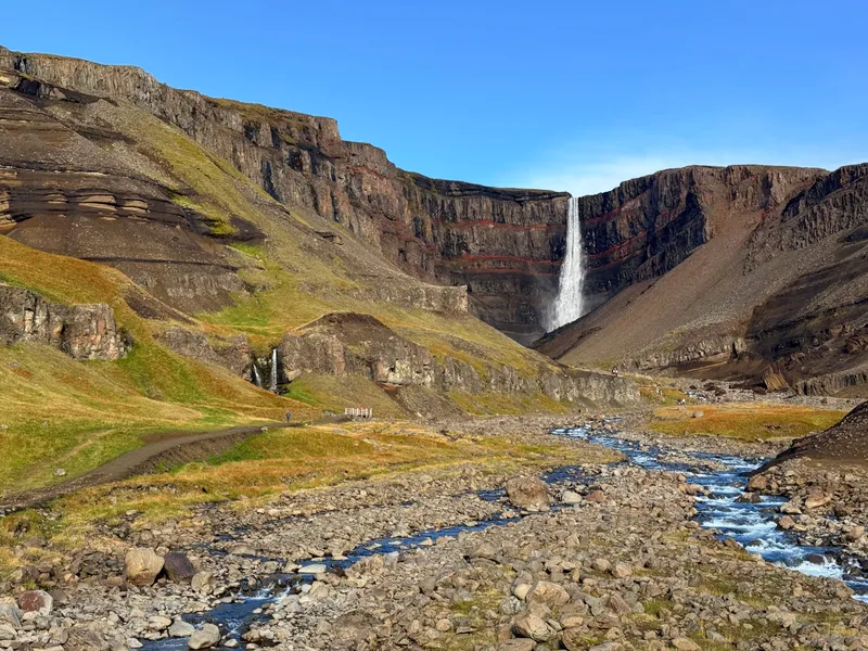 A tall thin waterfall falling into a banded cliff of dark basalt striped with bands of red clay, above a wide stony river valley in autumn colours