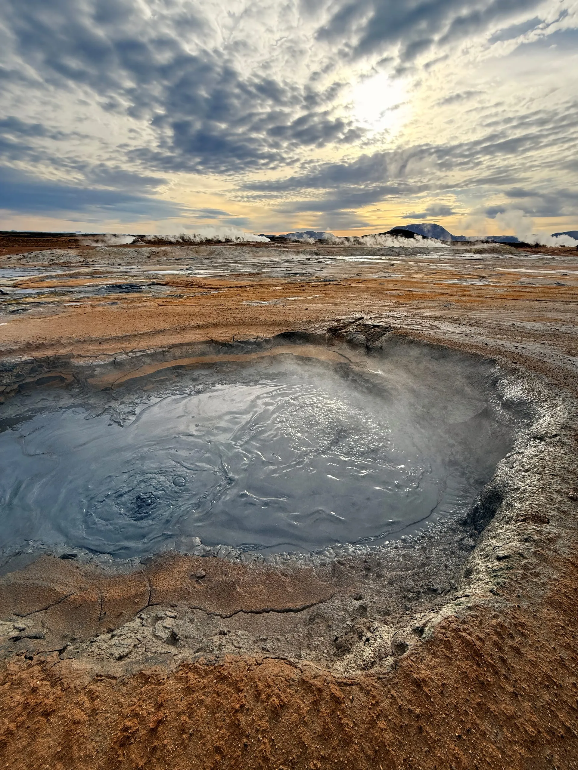 A grey mud pool steaming in a rust-orange geothermal plain under a mackerel sky, with vents smoking along the ridge behind
