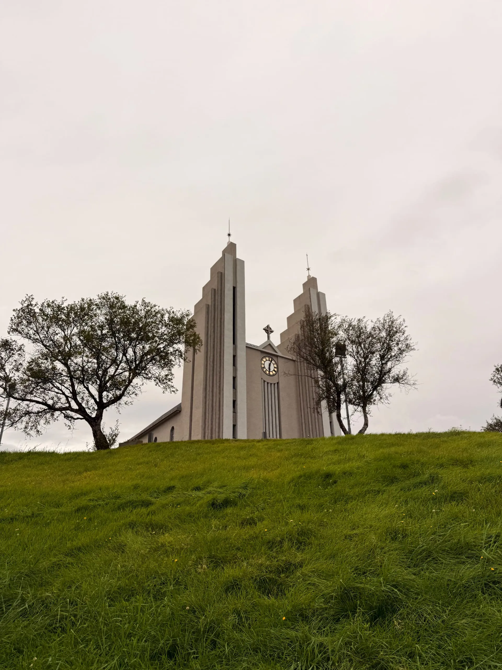 A pale concrete church with two stepped towers seen from below across a steep green bank, under a flat grey sky, with bare trees on either side